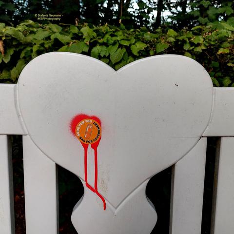 Decoration on the back of a white, wooden bench in the shape of a white heart. Some graffiti and a sticker make it look like it has been shot.