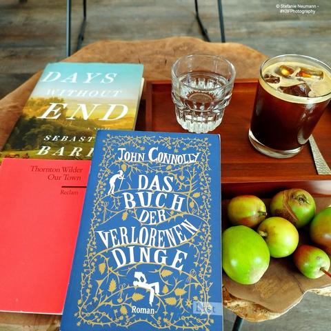 Ein kleiner Tisch in einem Café, voll gestellt mit einem kleinen Holztablet, welches ein Glas Kaffee auf Eis, ein Glas Wasser und einen Löffel enthält, sowie drei Büchern und sechs kleinen Äpfeln.
A small table in a café, set with a small wooden tray containing a glass of coffee on ice, a glass of water and a spoon, as well as three books and six small apples.