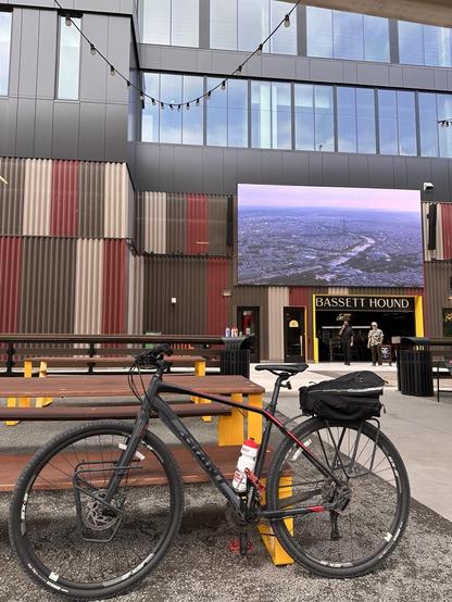 A black bicycle leans against an outdoor table. It is daytime. A darkened string of lights hangs above. There is a bar in the distance with the words “BASSET HOUND” above. A giant screen is mounted above the bar with a scene of Paris showing on it. 