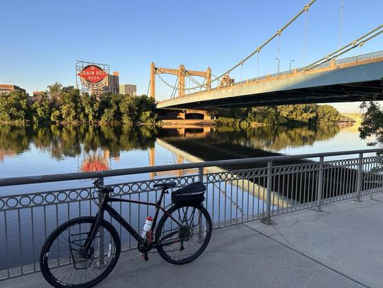 A black bicycle leans on a metal railing next to the Mississippi River. Sunlight lights up a bridge behind. Across the river is a round red sign that looks like a bottle cap. It has the words’Grain Belt Beer’ on it. The sky is clear and blue and the water is reflecting the blue color as well as the red sign and the bridge.