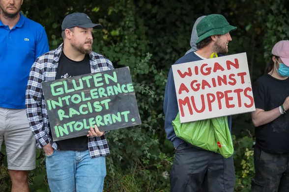 Picture from the antifacists, or counter protesters if you prefer, who went out on July 7th 2024 and stood up against the rancid far right terrorists. Pictured is a section of the crowd there are 4 people pictured from right to left a person in a pink hat, blue face mask, blue t-shit, a person in a green hat, dark blue shirt and grey trousers, holding a placard that says 'Wigan Against Muppets', a person in an open check shirt, black T-shirt, and blue Jeans with one hand in there pocket and a sign that says 'Gluten Intolerance, Racist Intolerant', and a person wearing a blue lacoste short and cream shorts...all are stood in front  of a green leafy hedge.