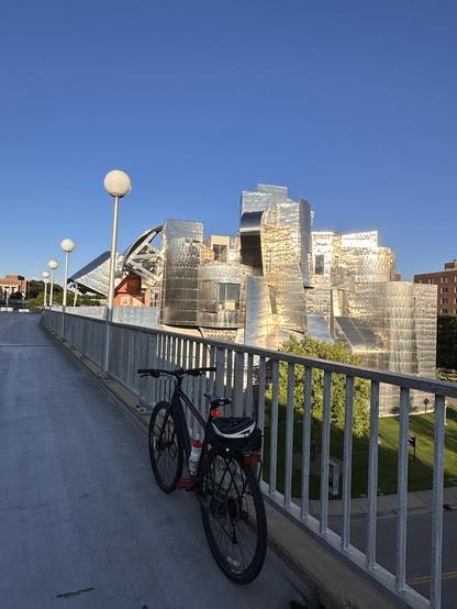 A black bicycle leans against a railing atop a bridge. In the distance a shiny, metal, modern building reflects sunlight.