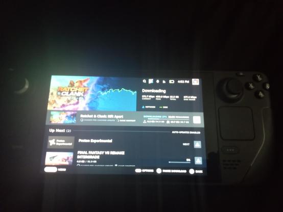 A picture of a Steam Deck on my bed in a rather dim light due to my camera being poor in lighting. On the Steam shows a rather blurry image of some games being installed.