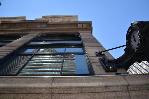 A buildings windows reflect another building. A black clock suspended at the corner of the building shows its 12 o’clock.Above is a blue sky.