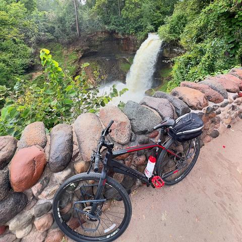 A black bicycle stands next to a rock wall. Beyond is a green, forested area with a waterfall.