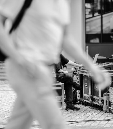 A grayscale photograph. A human figure filling most of the frame, out of focus, crosses from left to right.  In the space under one of that person's arms, we see part of another figure, working an audio board, framed.