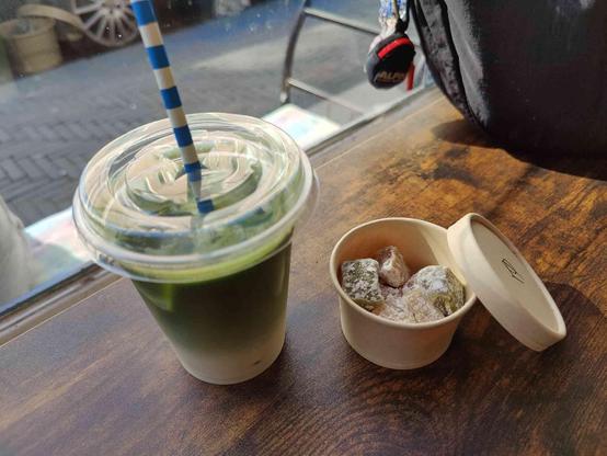 A photo of an iced matcha latte with oat milk and habutae mochi (traditional confectionery that originates from Fukui) in a small round cup with a lid. They are placed on a wooden table with my black bag from Uniqlo. In the background is the outside world and it is pretty sunny. 