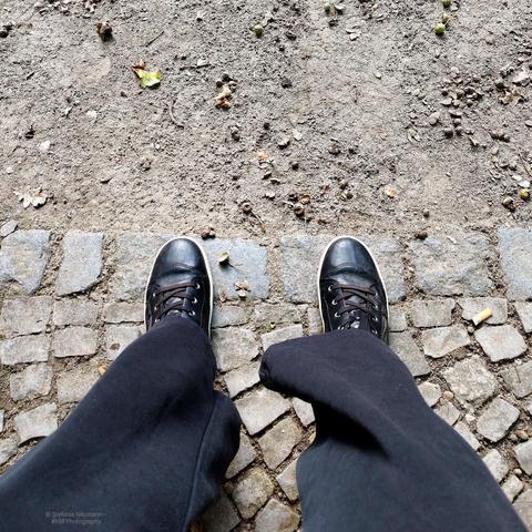 Two legs in black pants ending in black-shoe-d feet on cobblestone in front of acorn-covered sandy ground.