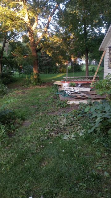 Looking into the backyard there are bare spots and weeds where debris and work stations have been. The deck has ladders, bits and bobs all over it.