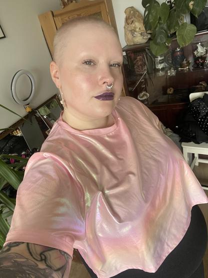 selfiessä lähes kalju tatuoitu henkilö päällään vaaleanpunainen hologrammi croptop. Nenässä iso septumkoru ja huulissa tummanviolettia kiiltävää huulipunaa.