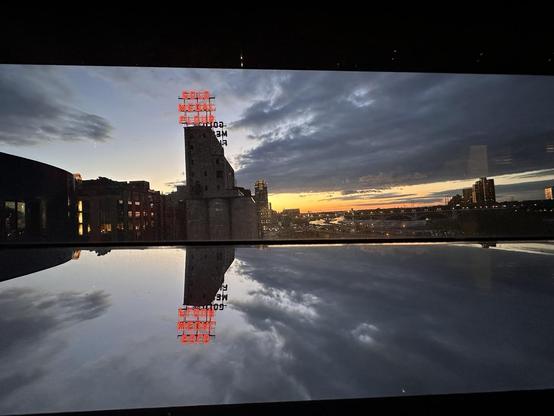 View through a window with a mirrored frame, of an old grain elevator and a darkening evening sky. Atop the building are neon red letters that read ’Gold Medal Flour’. The image is reflected in the mirrored window frame upside down. 