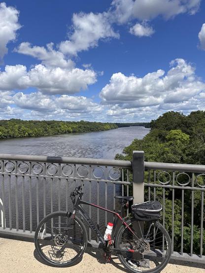 A black bicycle leans on a railing of a bridge looking over a view upriver of the Mississippi River. The river banks are forested and green. In a blue sky above cumulus clouds float by.