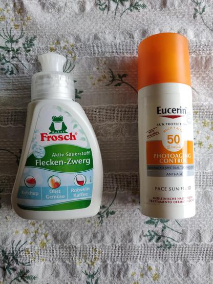 Auf einem Tisch liegt links der Frosch Flecken-Zwerg für Ketchup, Obst & Gemüse, Rotwein & Kaffee, rechts liegt eine kleine Dose mit Eucerin Photoaging Control Sonnenschutz.