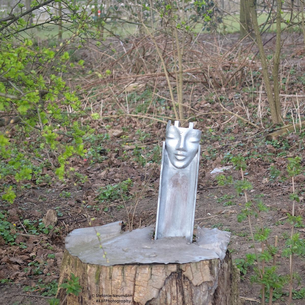 Eine kleine Statue aus Metall auf einem alten Baumstumpf installiert. Die Oberfläche des Stumpfes ist mit Metall beschlagen und darauf senkrecht eine kurze, metallene Schiene angebracht, in die oben ein Gesicht eingearbeitet ist. Die Farbe des Metalls <br />ist silber. A small metal statue installed on an old tree stump. The surface of the stump is covered with metal and a short, metal rail is mounted vertically on it, with a face moulded into the top. The colour of the metal is silver.