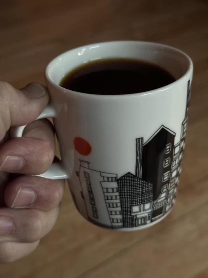 A mug of coffee is white and a a scene of silhouetted homes on it with a round orange sun rising.