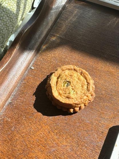 A round cookie with a textured surface is placed on a wooden tray, illuminated by sunlight, casting a soft shadow.