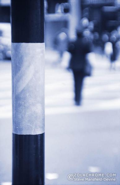 Monochrome photograph of a city street show a striped pole in the foreground and an out of focus image of a person in the background.