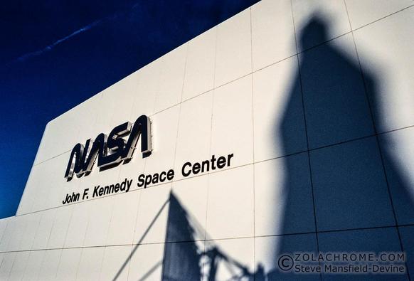 Front wall of a white building against a blue sky with the sign 'NASA John F. Kennedy Space Center' with large shadows of a rocket and a satellite dish.