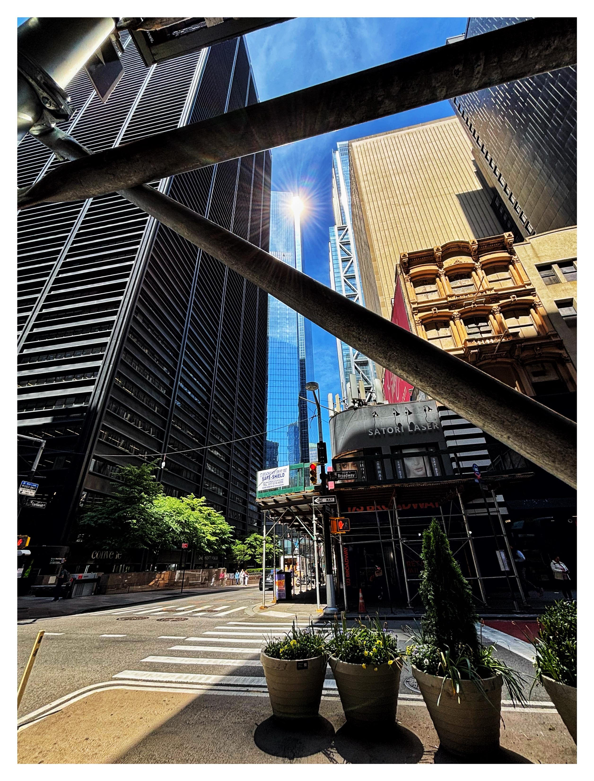 Wide-angle view of a city block in lower Manhattan, dominated by a starburst of sunlight reflected from the top of a distant glass skyscraper. The starburst is framed by two scaffolding bars close to camera, which intersect to suggest a “<“ shape or triangle. In the middle ground are high-rise buildings and an older low-rise building with its facade obscured by scaffolding and Italianate windows on its upper floors. In the foreground are large potted plants on the median of a street intersection. Bright sunlight illuminates the building facades and sidewalk trees, while a building shadow divides the street surface into sunny and shaded areas. The slice of sky beyond the buildings and scaffolding bars is a vivid blue with wispy white clouds.