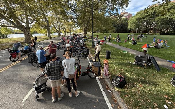 Memorial Drive in Cambridge, MA, facing east with no cars on it because it is Sunday afternoon.  There's a long line of cargo bikes parked in one lane, with people standing in and around them.  JFK park is on the right, with a bunch of kids playing with stuff provided for their amusement (a giant tetris yty, some bubbles).

I got lucky and not too many faces are visible, I think the ones that are, were associated with the event.  I took the photo standing on the deck of my cargo bike, as one …