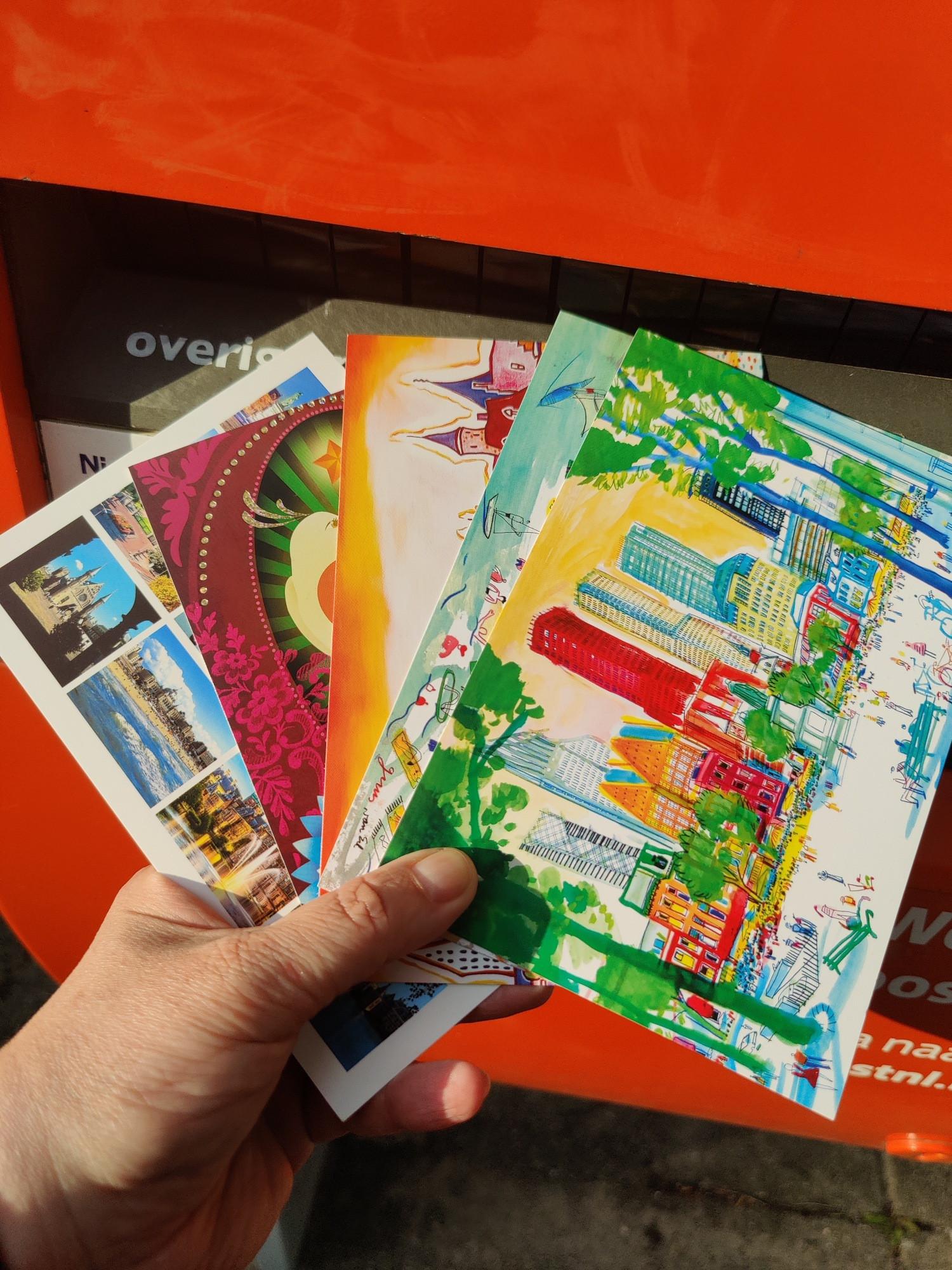 Five cards with photos or drawings of The Hague, the beach, and a card with birds. They are about to be dropped in the red post box of PostNL.
