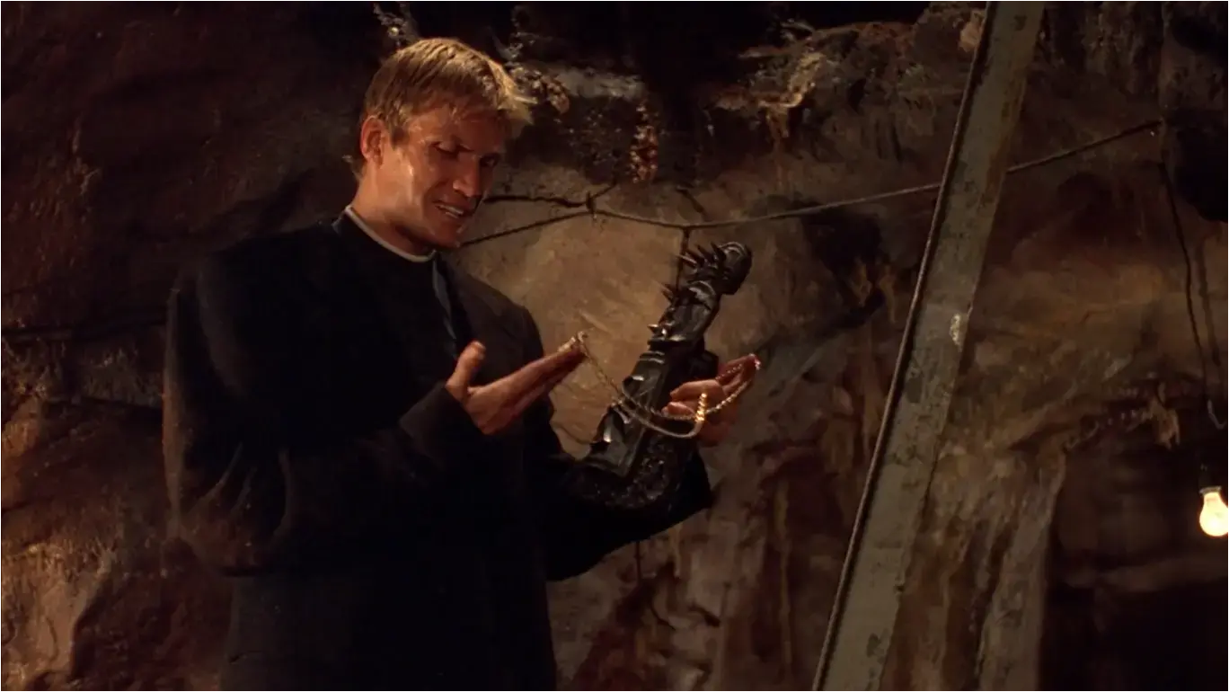 Screenshot from the film &#39;The Minion&#39; (2016) - Dolph Lundgren in priest&#39;s garb in a poorly-lit underground tunnel, looking at a golden key on a chain he&#39;s holding, whilst also holding a spiked fighting glove