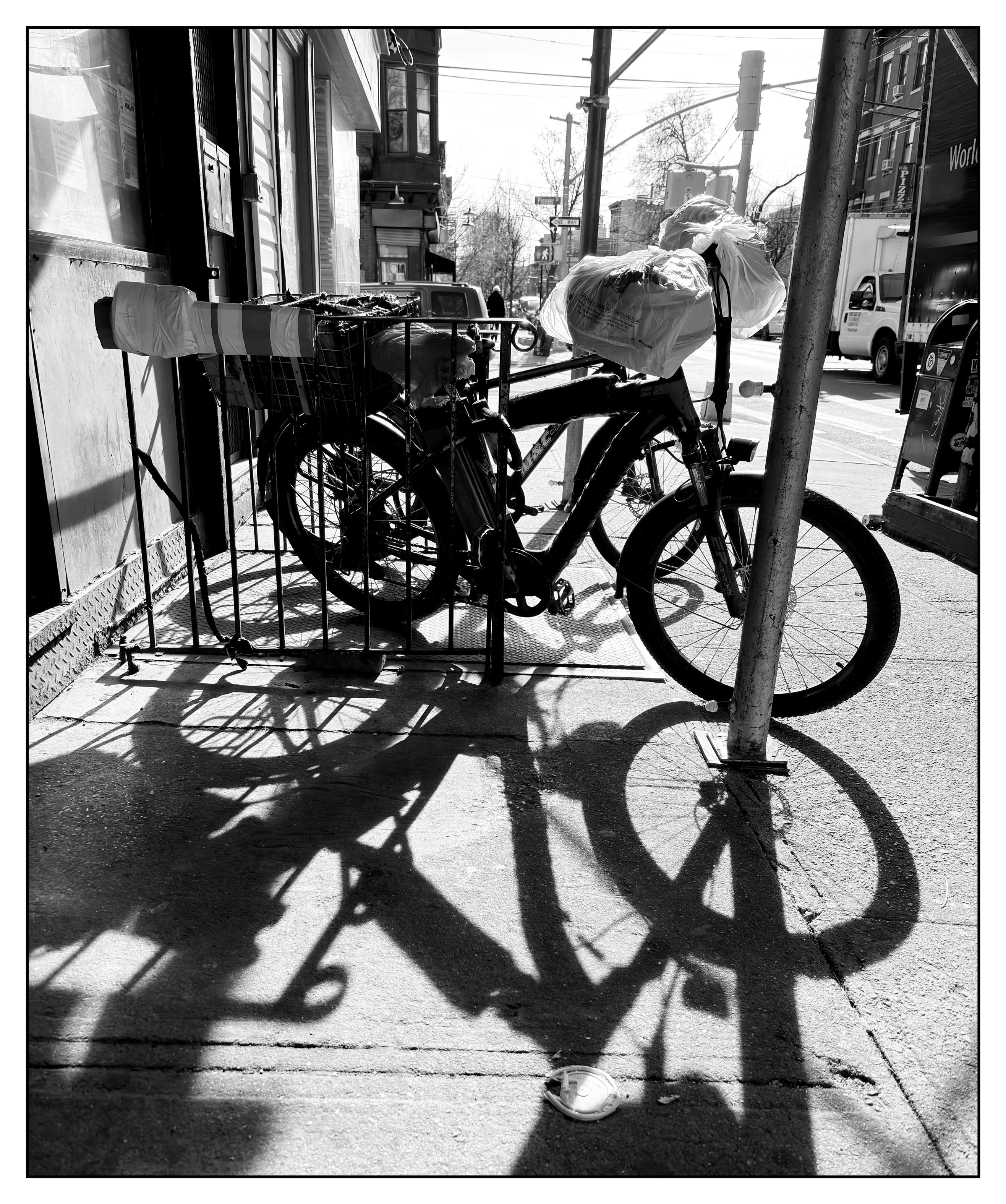 Black and white photo of a parked bicycle on a city sidewalk, backlit by the sun. Its wheels cast dark shadows into the foreground, making them resemble figure eights.