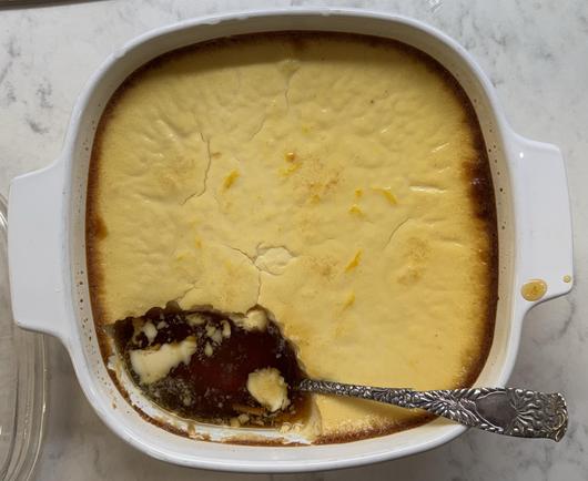 A ceramic dish containing a flan with a serving removed from its lower left, showing the yummy brown caramel juice.  An ornate sterling silver serving spoon is in the dish with its bowl resting in the caramel juice and its handle extending out of the lower right corner of the dish.