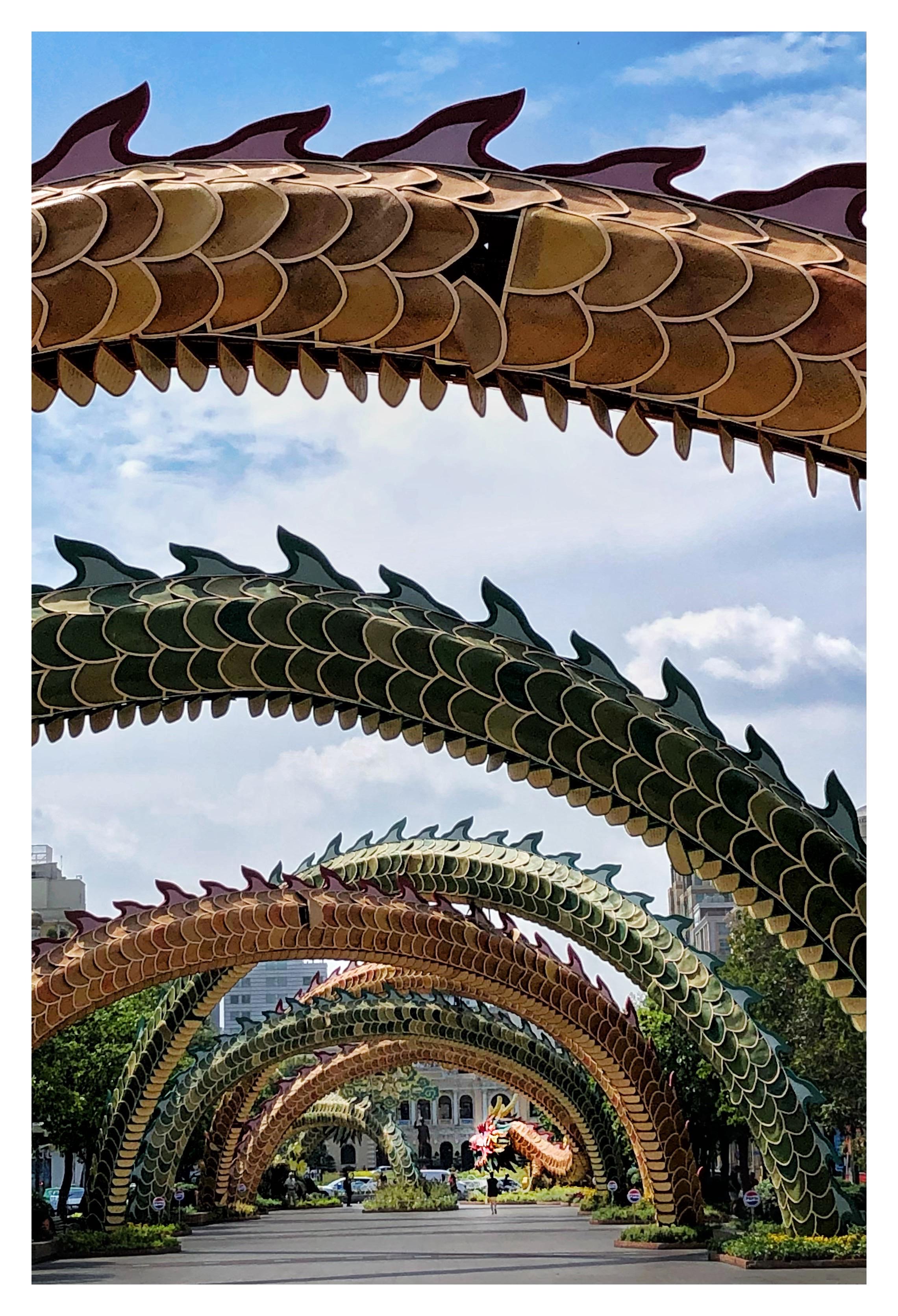 Photo of a series of swooping arches over a wide pedestrian avenue receding to a building in the distance. The arches have scales and fins in hues of orange and green, and the arch at the far end bears a sculpted dragon’s head. The effect is of a dragon’s long body encircling the avenue, leading the eye to a statue of a man with one arm raised (Vietnamese leader Ho Chi Minh) in front of a palatial facade. Trees and shrubs line the sides of the avenue, and the sky seen through the dragon arches is blue with white clouds.