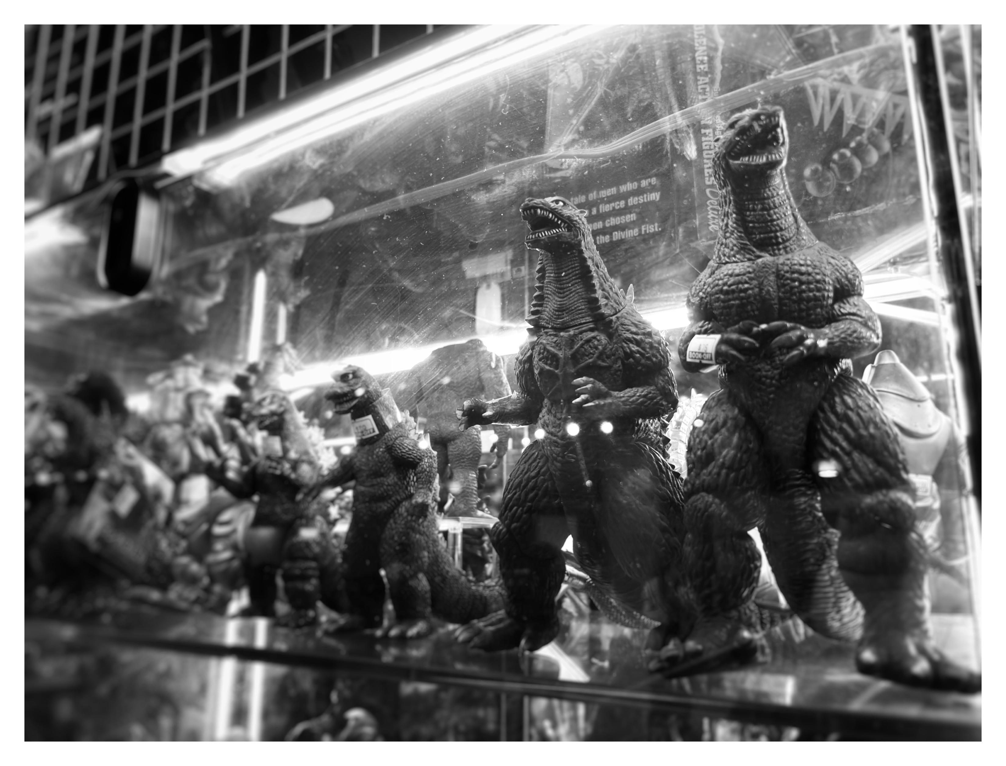 Black and white photo of several miniature Godzilla figures of different heights, arranged in a row in a glass store case. The photo is angled so that two taller Godzilla figures on the right are closer to the camera and in focus. The toys are backlit by fluorescent lighting that shows some of the scratches on the case. Photographed at Japan Village in Brooklyn.