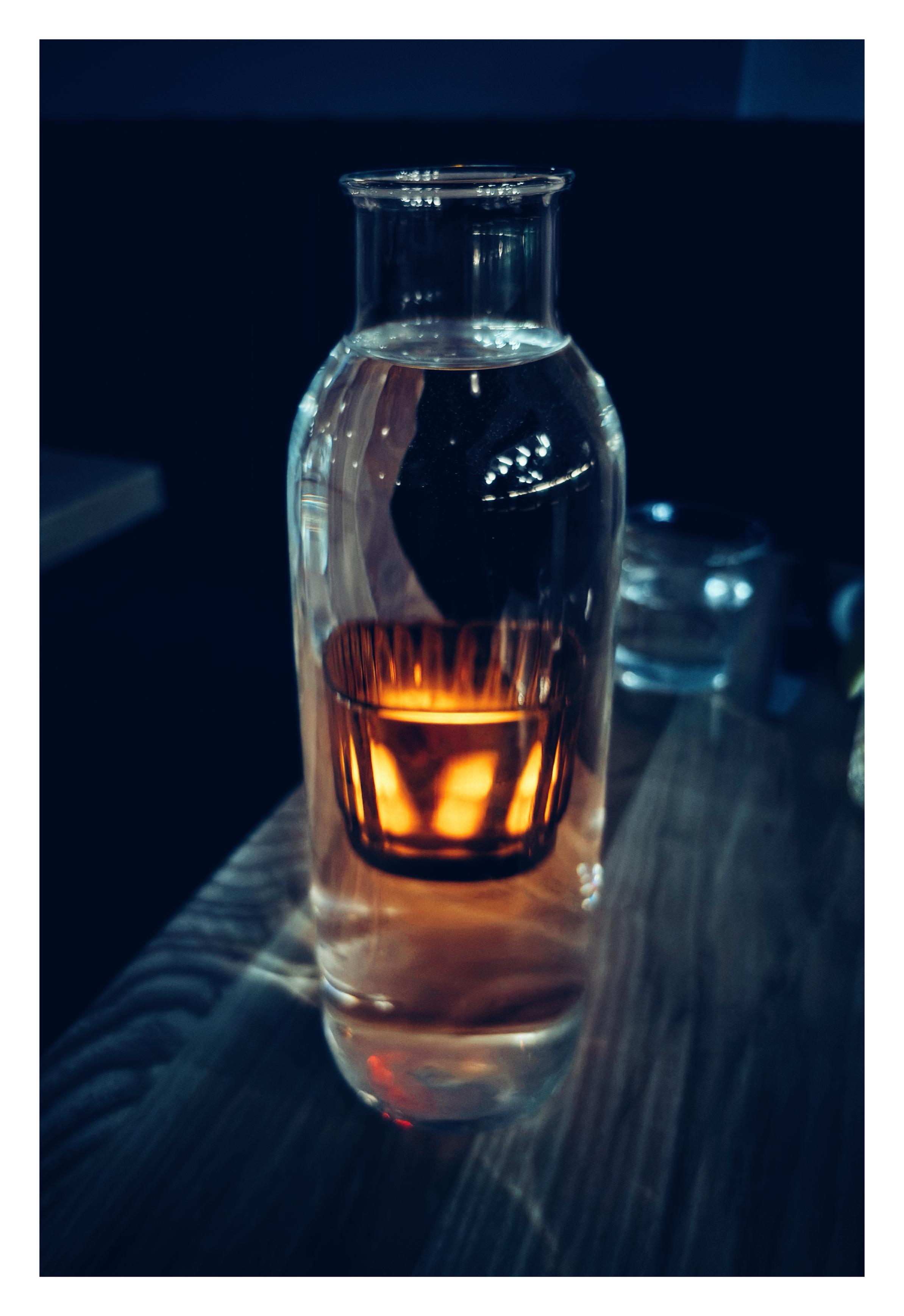 A tall carafe filled with water sits on a restaurant table with wood grain. Through the water we can see a refracted image of a small four-sided glass tumbler sitting directly behind the carafe; the tumbler’s sides glow with a warm yellow light, illuminated by an electric candle inside. The tumbler appears to float inside the carafe, levitating above the table. A blurred drinking glass can be seen farther away on the table, and the rest of the background is in cool blue-toned shadow.