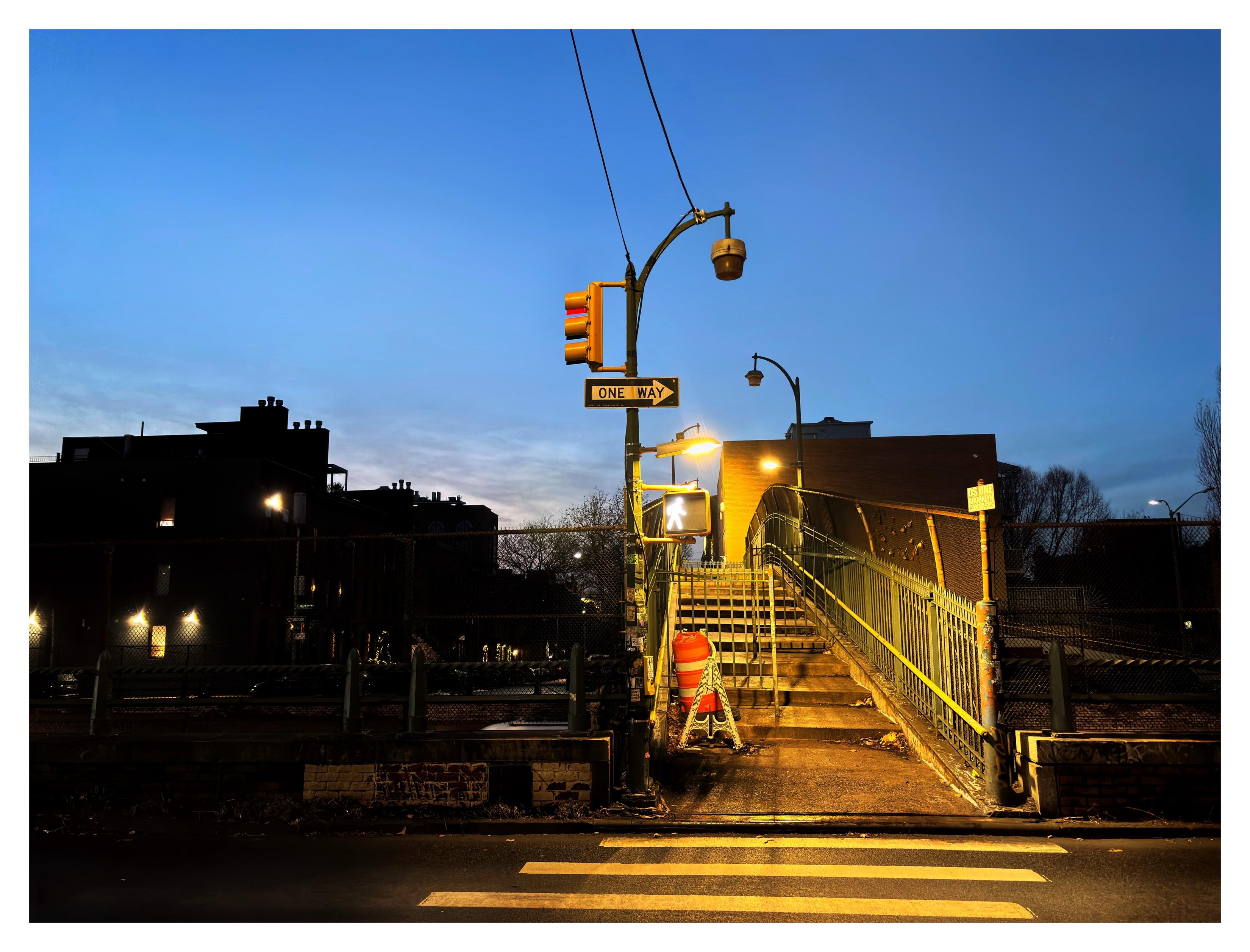 A pedestrian bridge an hour past sunset, under a deepening blue sky. The stripes of the crosswalk in the foreground are echoed by the wide steps on the ascending part of the bridge. On one of the steps sits a cylindrical orange and white traffic barrier. At the entrance to the bridge is a streetlamp with several things attached to its pole: a traffic light, a “One Way” arrow sign, and a pedestrian signal with the “walk” symbol lit up. Two cables extend from the streetlamp’s arm up out of the photo’s top edge. Lights over the bridge cast a warm orangey glow. In the distance are shadowy low-rise buildings with some lit windows. That’s a lot of description for what’s really a pretty nondescript view; I just liked how it was lit in the blue hour.