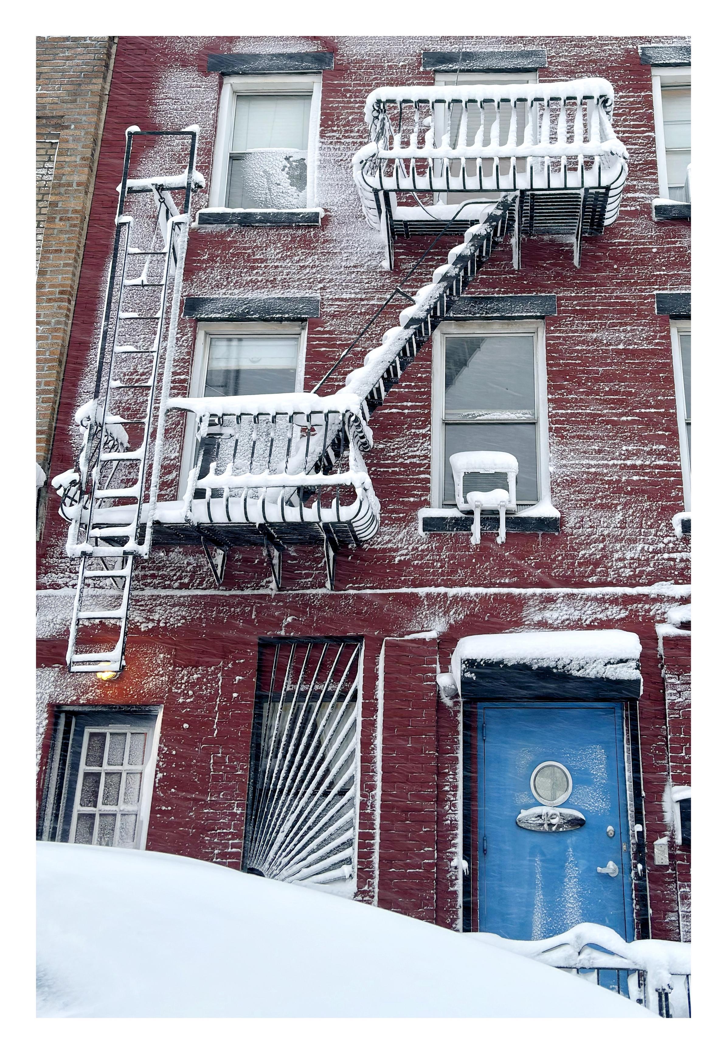 The red brick facade of a low-rise apartment building in the middle of a blizzard. Snow clings to the walls and thickly covers all upper surfaces โ windowsills, door lintels, the ladders and railings of fire escapes. At street level is a blue door with a round glass portal. Beside it is a tall narrow window with a design of metal bars radiating out from its lower left corner. Partly obscuring the door and window is a mound of snow in the foreground, hiding a buried car. If you look closely you can see windblown snowflakes streaking all across the image.