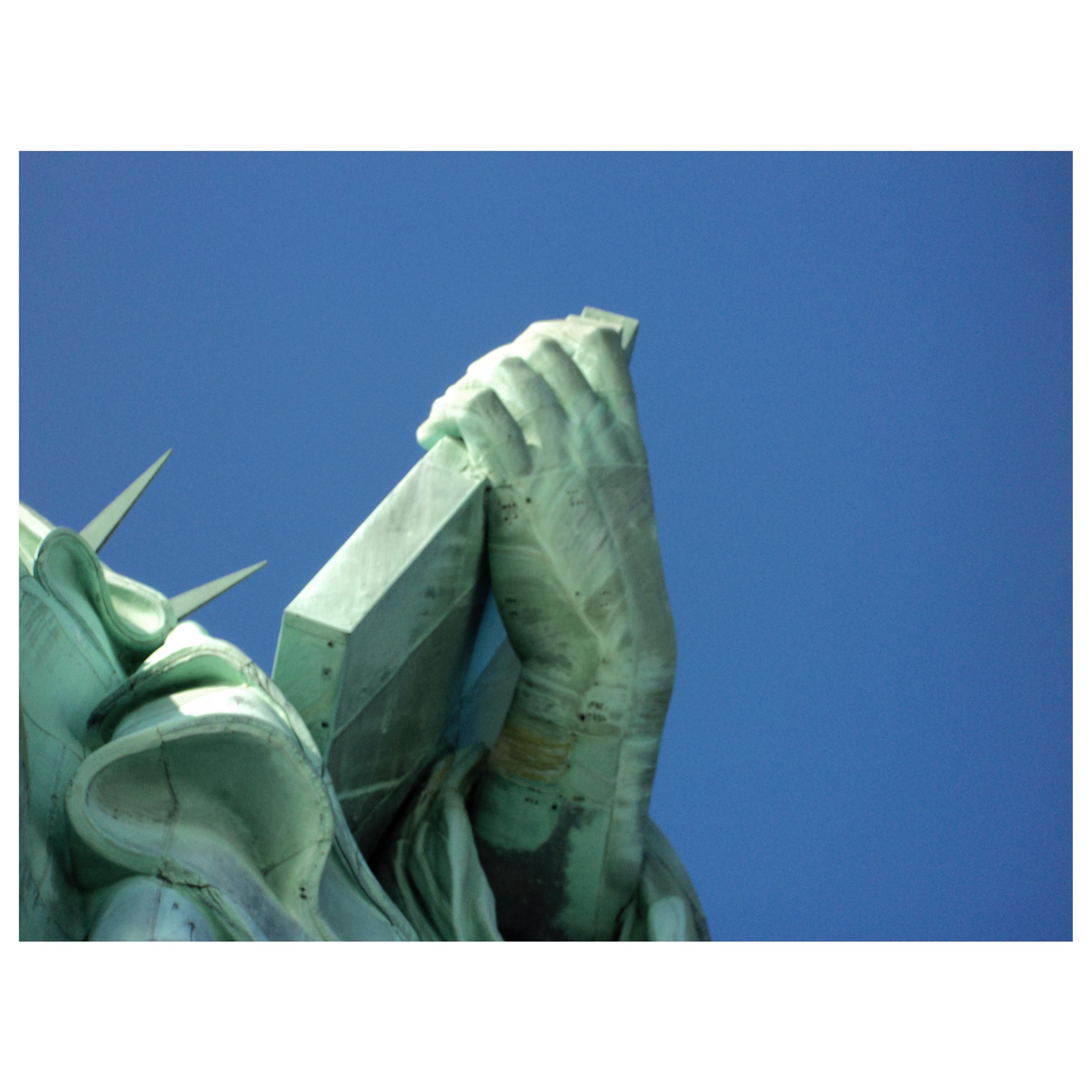 A view from almost directly underneath the Statue of Liberty, looking straight up at her left hand holding the tablet. We can also see a portion of her robes and two of the pointed spikes on her crown, all against a clear blue sky. The image features details such as the green hue of the statue’s weathered surface and the seams where the copper sheets have been joined together. I took this photo more than a decade ago during my last trip to Liberty Island.