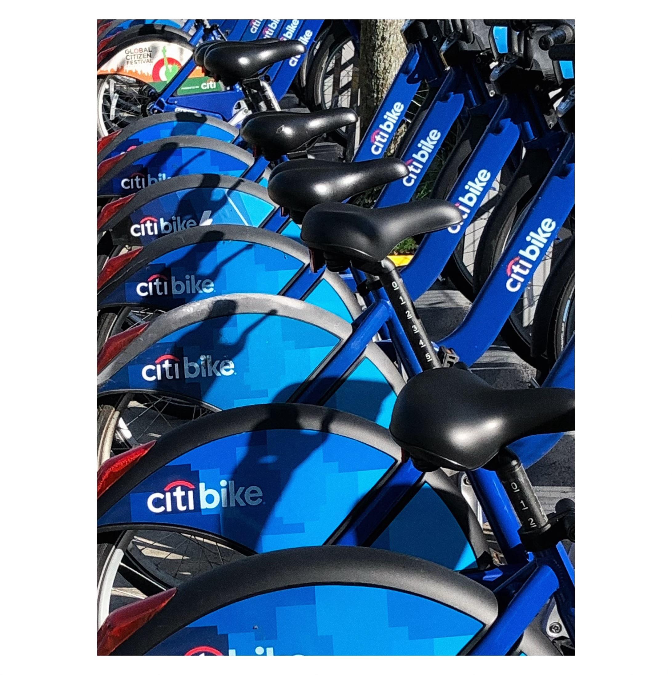 Closeup on the back wheels and seats of a row of blue NYC CitiBikes parked at their docking station. The bikes are viewed from an angle such that they line up diagonally behind each other and recede from camera. Most of the wheels are partially covered by blue advertising discs displaying the CitiBike logo, except for a bike at the far upper left which has a colorful disc promoting the Global Citizen Festival.