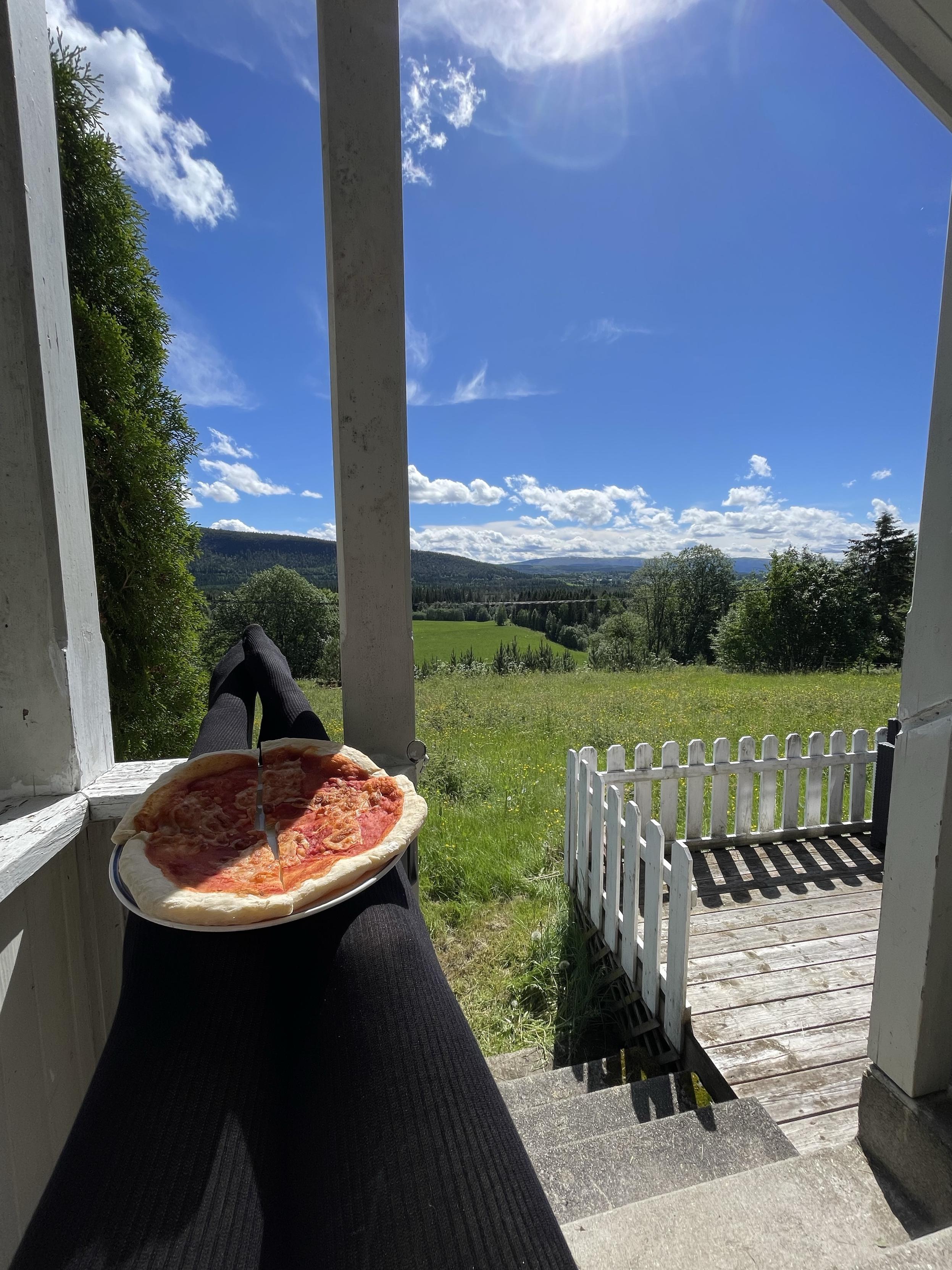 I am sitting on a porch with my legs crossed on top of a railing, holding a pizza on a plate. The background features a sunny landscape with green fields, trees, and a blue sky with clouds. A white picket fence and wooden steps can also be seen.