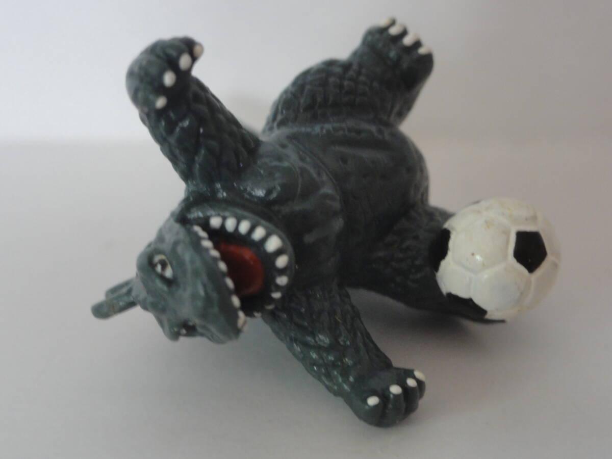Plastic keychain fob featuring Godzilla kicking a football. Lying on his back face up.