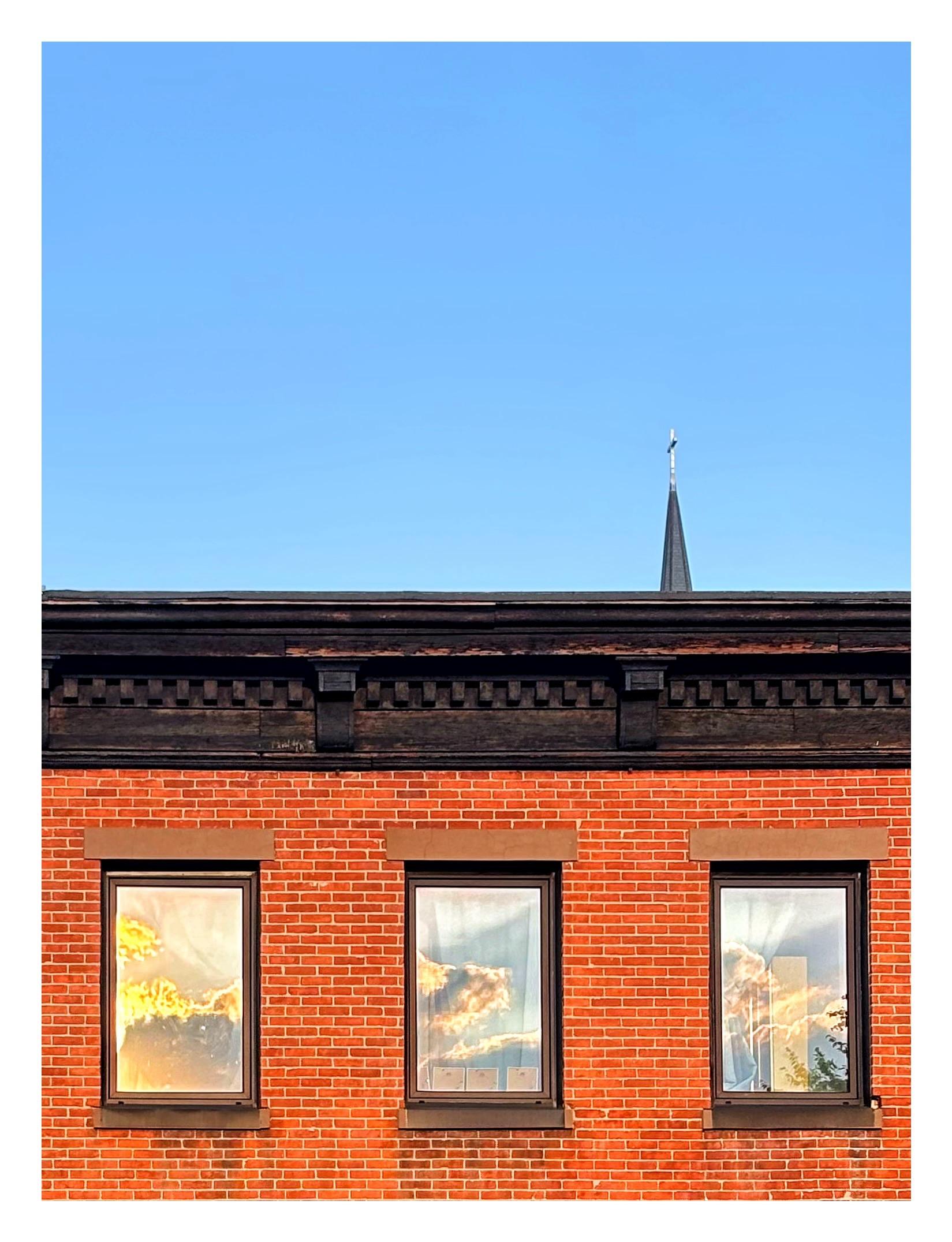 Three windows on the upper floor of a brick apartment building with a decorative cornice along its roofline. The steeple of a distant church pokes up above the roof. The windows face west and reflect cumulus clouds backlit by a golden sunset, while the eastern sky above the building remains clear and blue.
