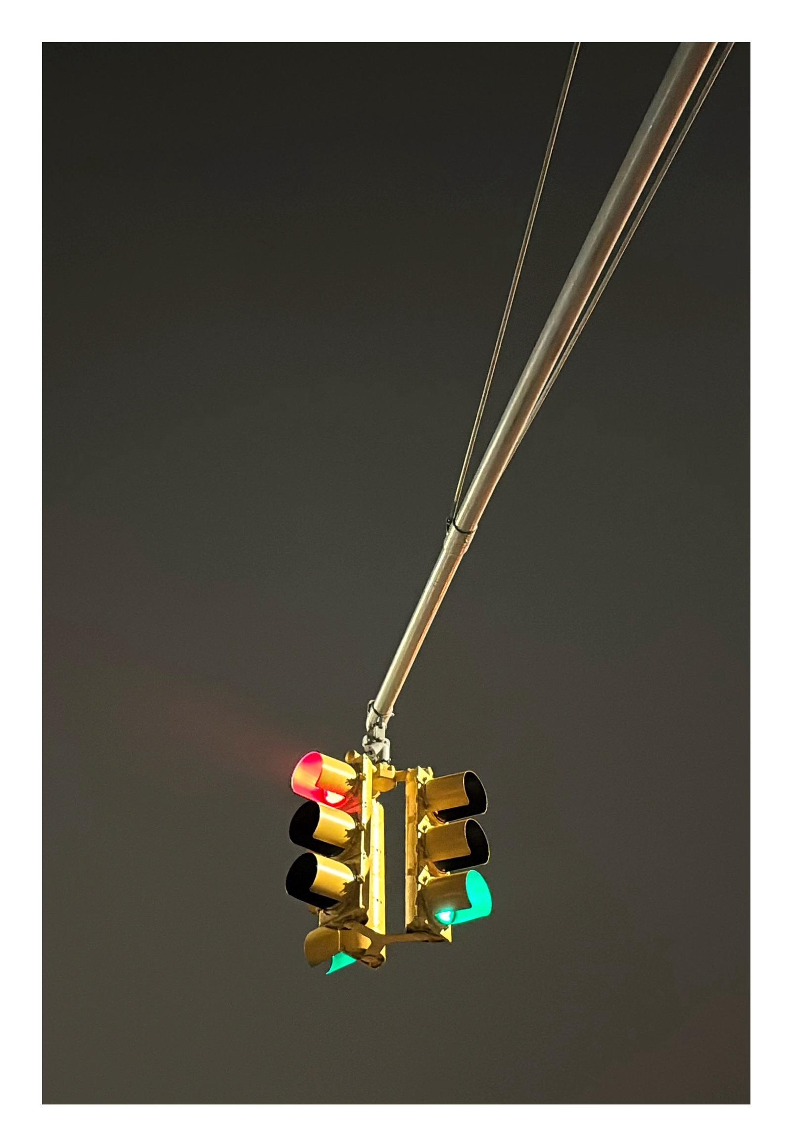Nighttime photo on a Brooklyn street corner, looking up. A traffic pole’s metal arm extends from the upper right of the image to the lower center, where a three-way traffic signal hangs, with two lights on green and one light on red. The sky in the background is a solid, featureless wall of gray.