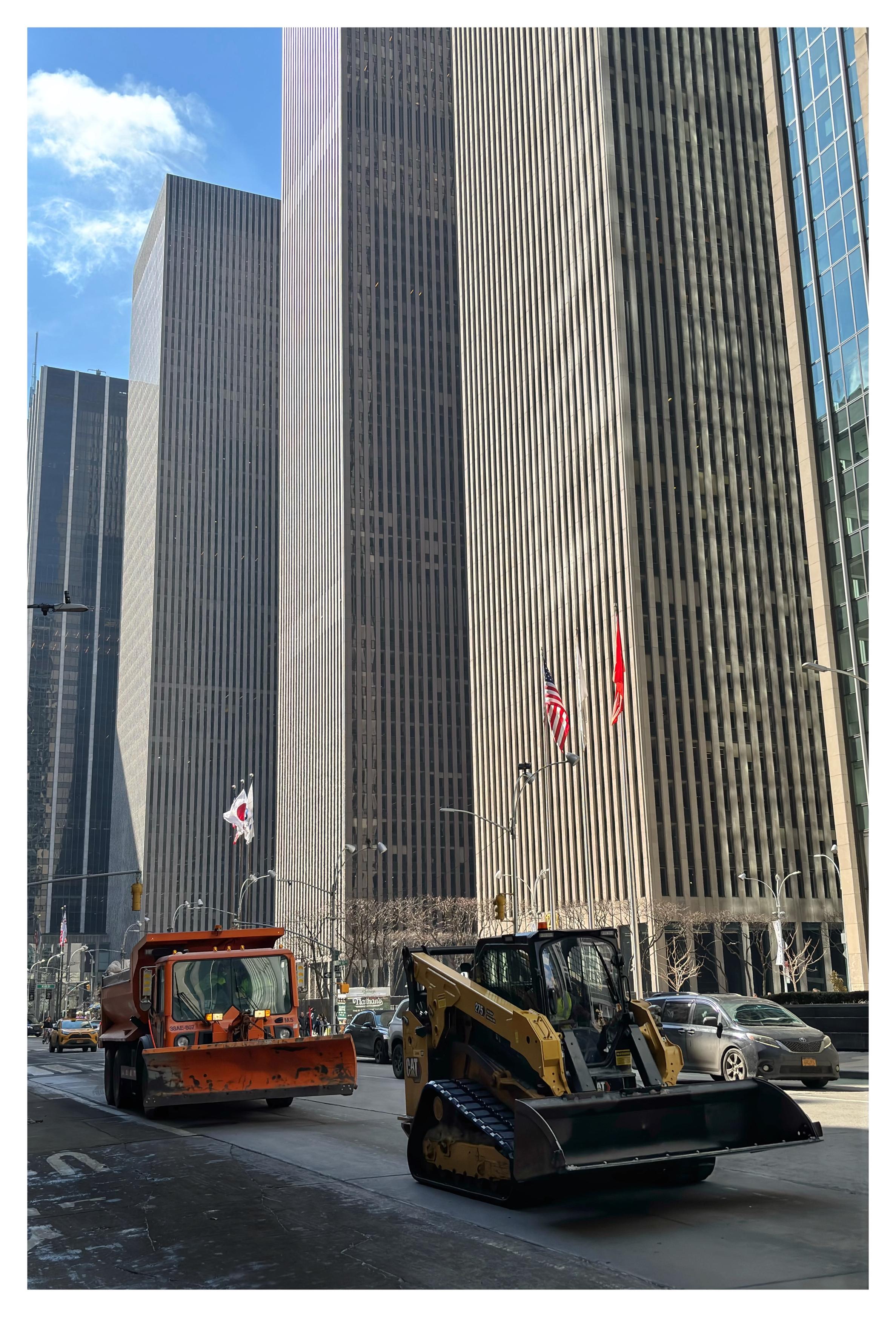 Two snowplows, one yellow and the other orange, chug along Sixth Avenue in Manhattan. There’s no trace of snow, even though a blizzard had hit the city a few days earlier. On the far side of the avenue are soaring skyscrapers. A bit of blue sky with white clouds can be seen at upper left.