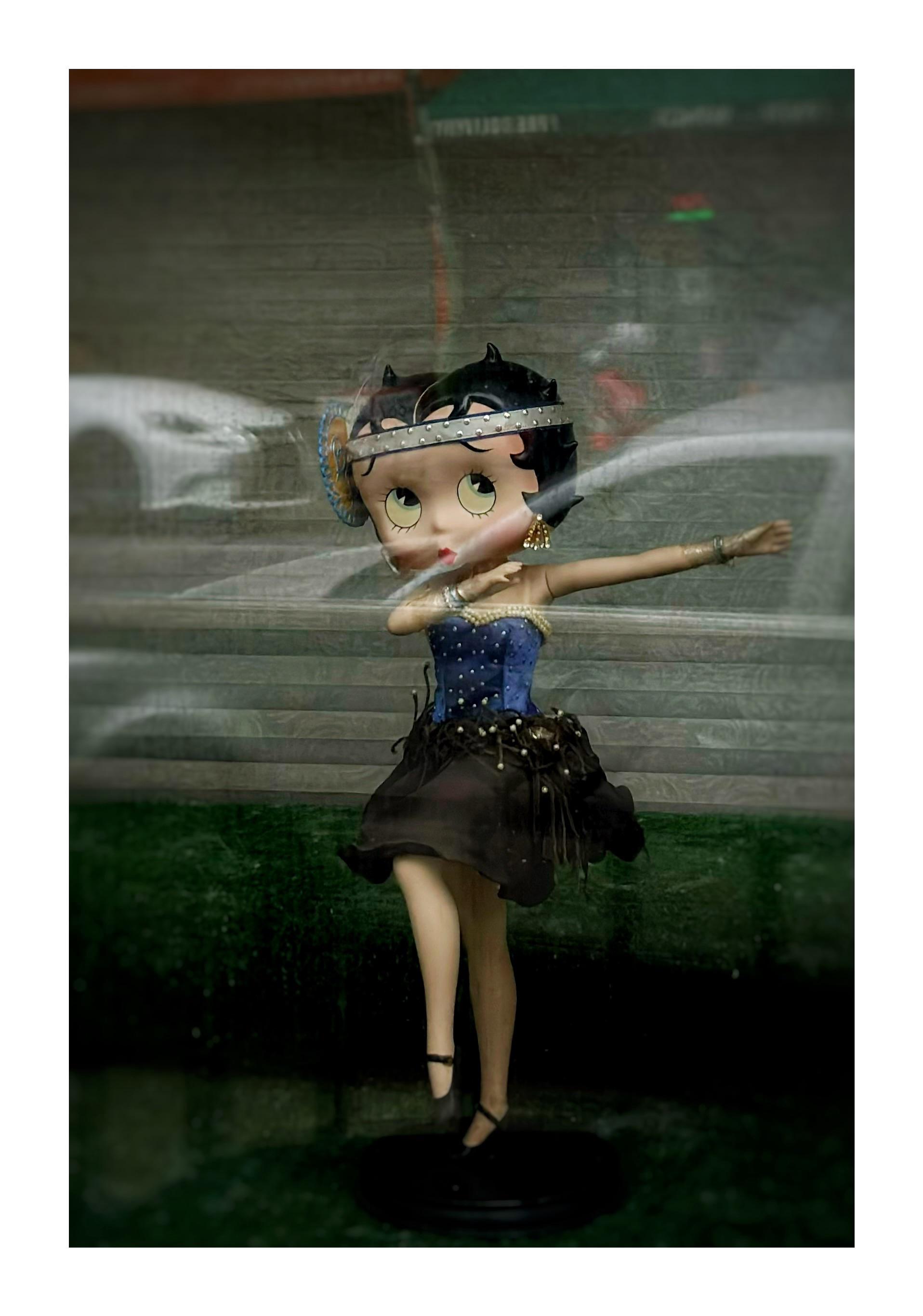 Photo of a Betty Boop figurine, one of several Betty Boops in a quirky window display in an apartment in Red Hook, Brooklyn. This one has been posed in a dancing position almost like a dap, with one arm extended and the other bent at the elbow, and with one leg raised. She’s wearing what looks like a 1920s flapper outfit: short black skirt, blue corset studded with pearls, silver bracelets, gold earrings, and a silver jewel-studded headband with a large blue-rimmed disc to one side. The figurine stands on a surface with a little step behind, all covered in green felt. The window glass reflects some cars parked at the curb and a storefront across the street.