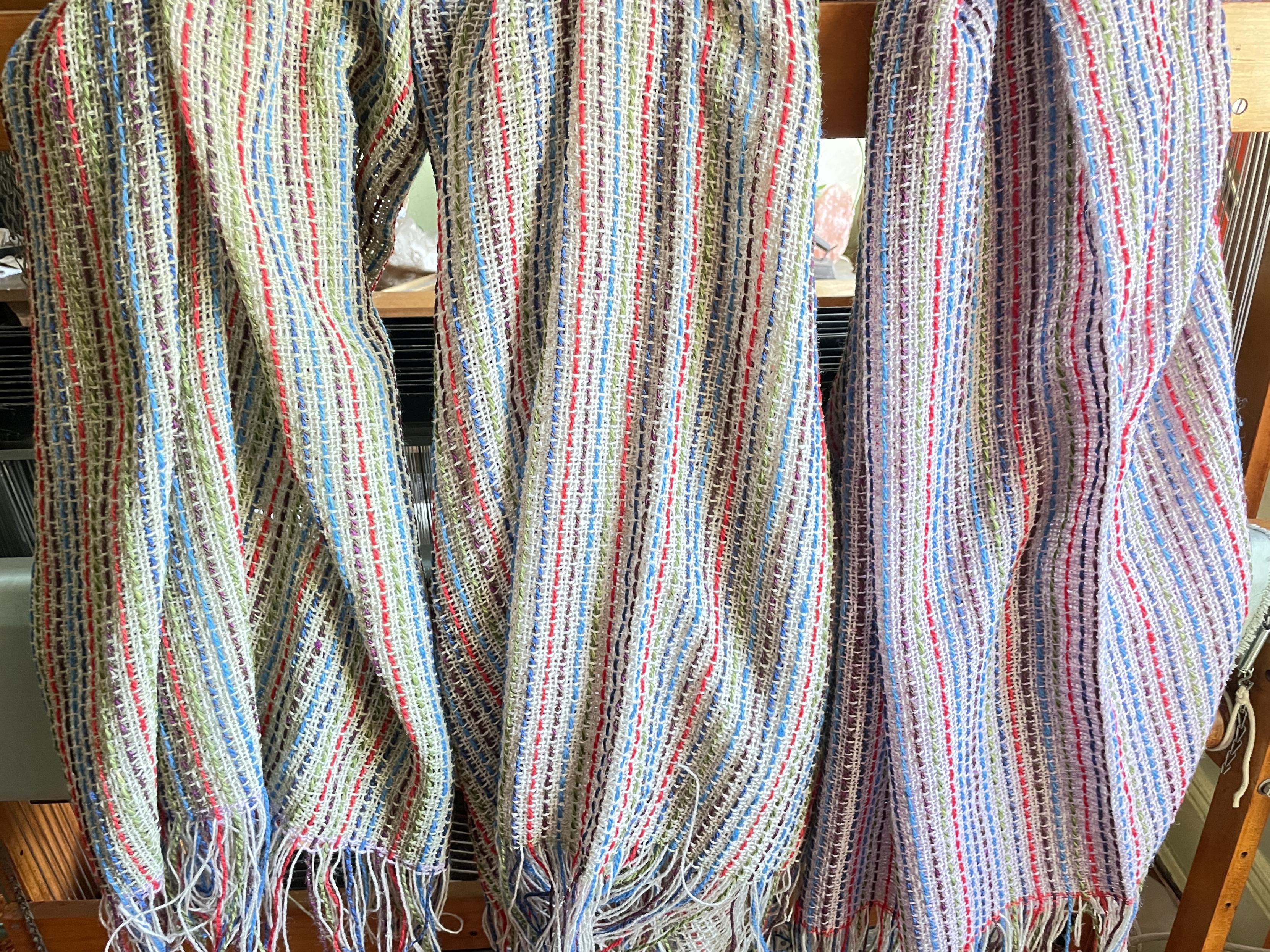 Photo of 3 handwoven blankets hanging.  They have lots of thin stripes of multiple colors on a lighter background.  The background of the one on the left is pale green, the middle is a light neutral and the one on the right is a pale lilac