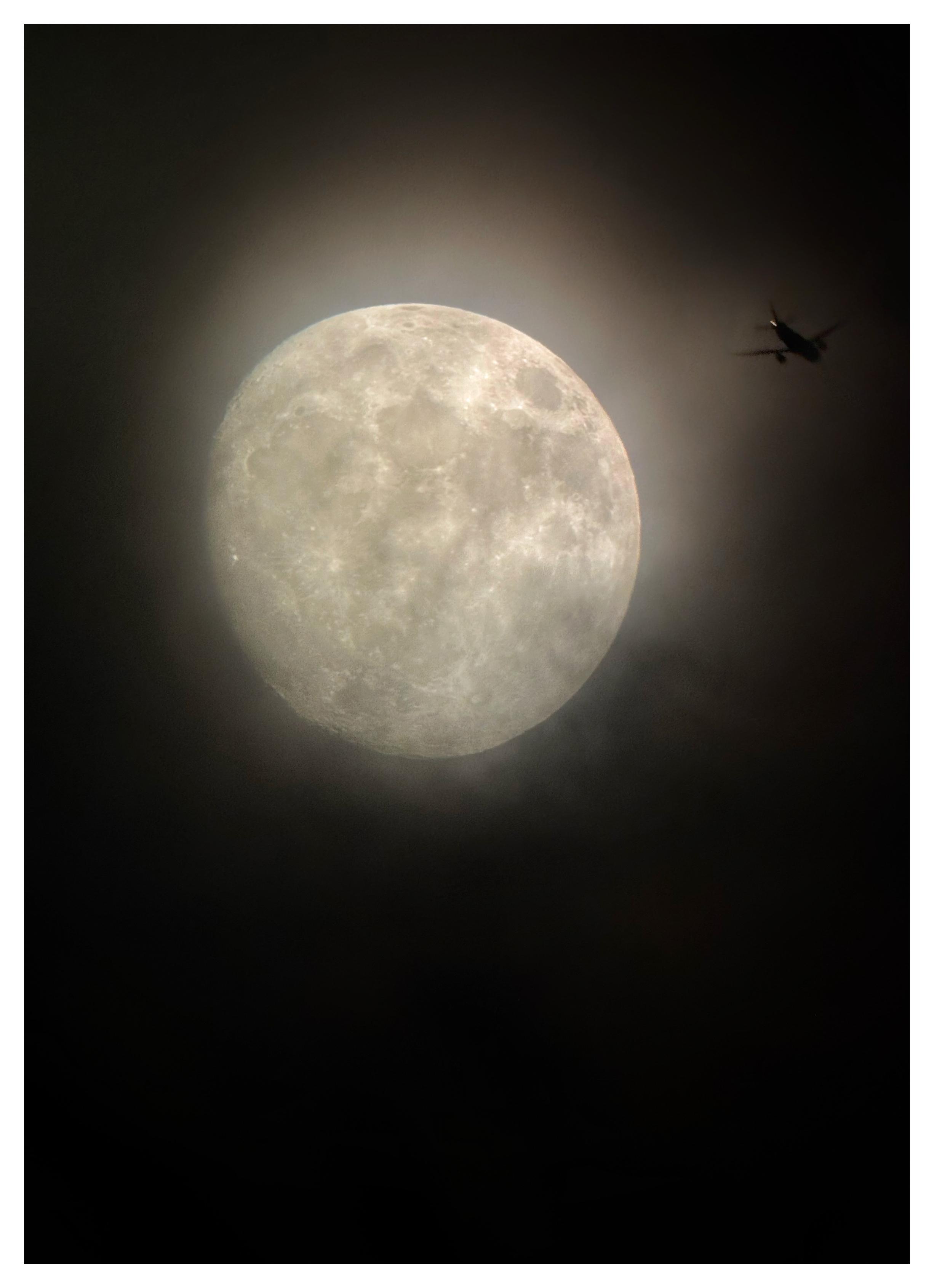 A waxing gibbous moon a few days before full, with visible craters. It casts a brownish glow against a dark sky, illuminating the shadowy edges of some clouds below. To its right is a small airplane, silhouetted against the moonglow, traveling rightward and away from the viewer.

Shot on an iPhone 15 attached to a Celestron 130mm-aperture reflector telescope.