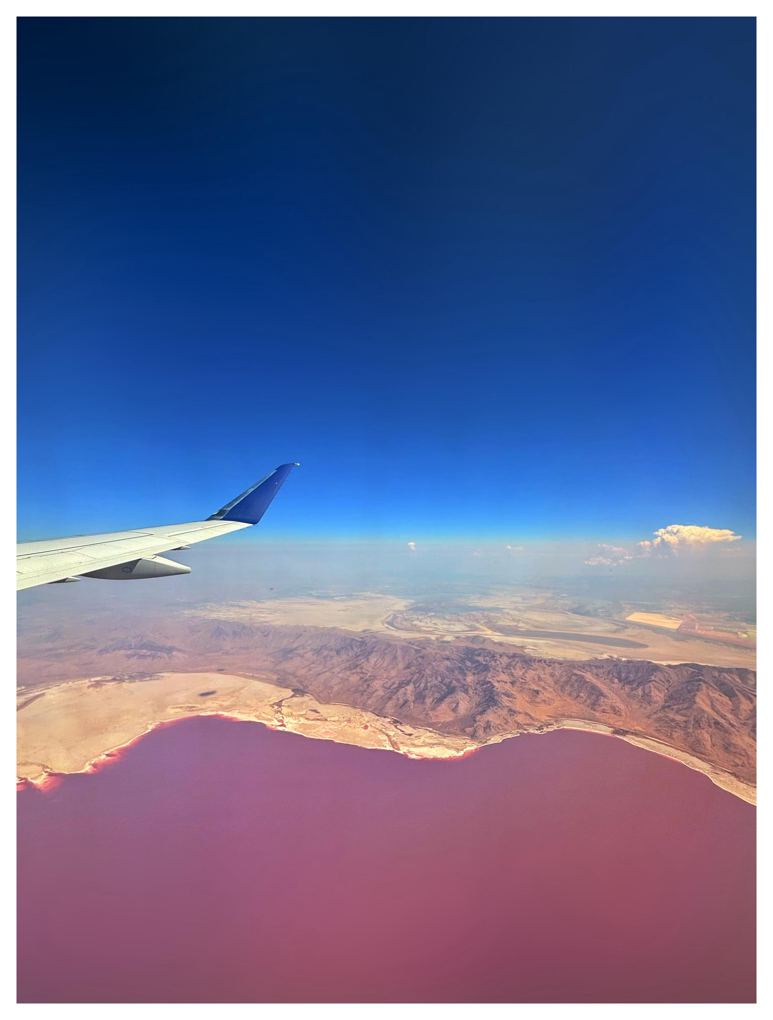 View of Utah’s Great Salt Lake from an airplane window. A brown, mountainous landscape sits between the wavy shoreline of a vivid red lake below and a clear blue sky above, with a distant cumulus cloud floating on the horizon. The airplane’s wing pushes in from left frame. The lake’s red color is caused by the pigmentation of halophilic (salt-loving) bacteria, which thrive in the high salinity of the water.
