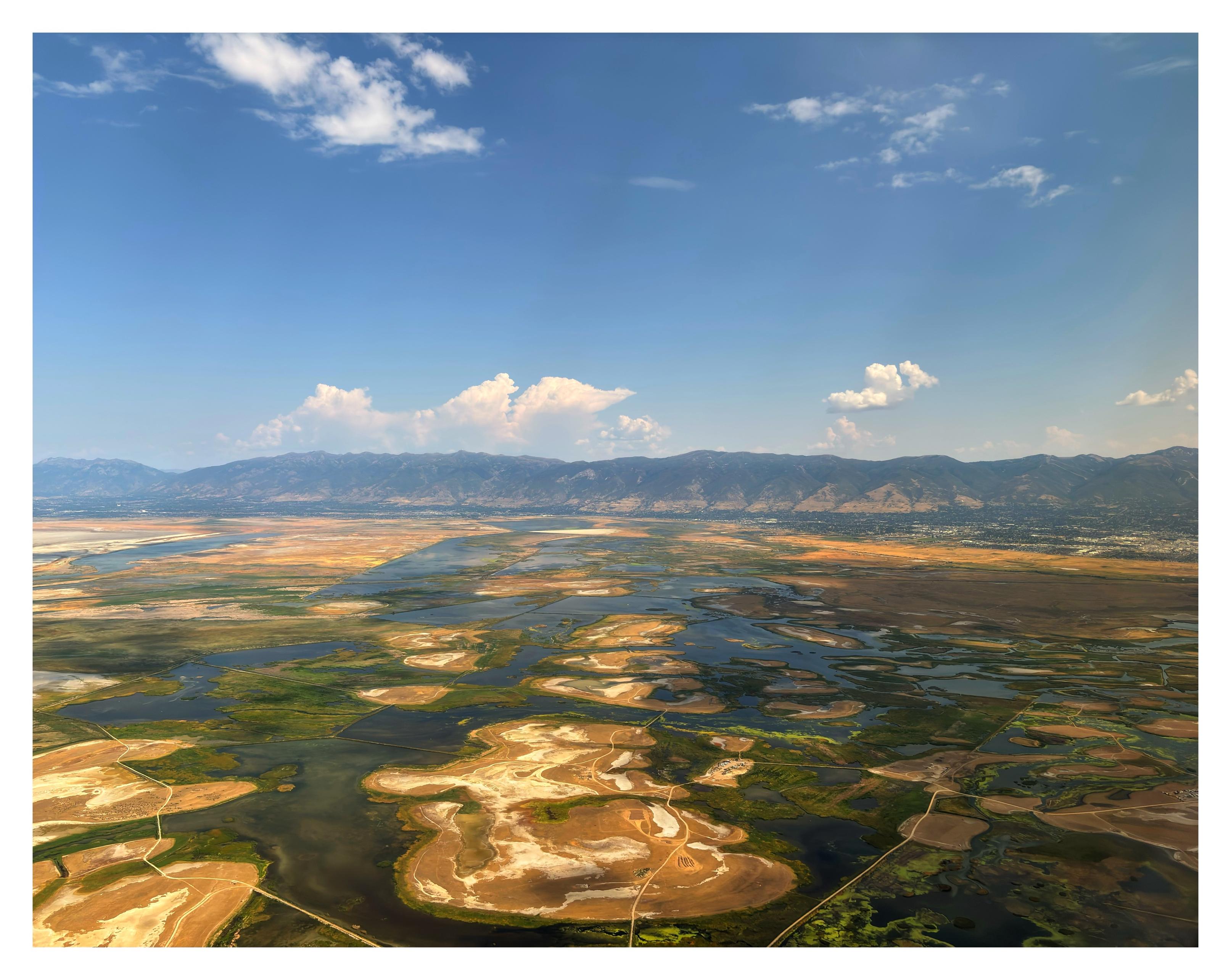 Another view of a Utah landscape from an airplane window. Flat islands of green and brown, connected by a web of narrow causeways, sit in what looks like waterways or a collection of small rivers and lakes — or perhaps these are the drying-up remnants of a larger body of water. Mountains sit at the horizon under a blue sky with clouds.