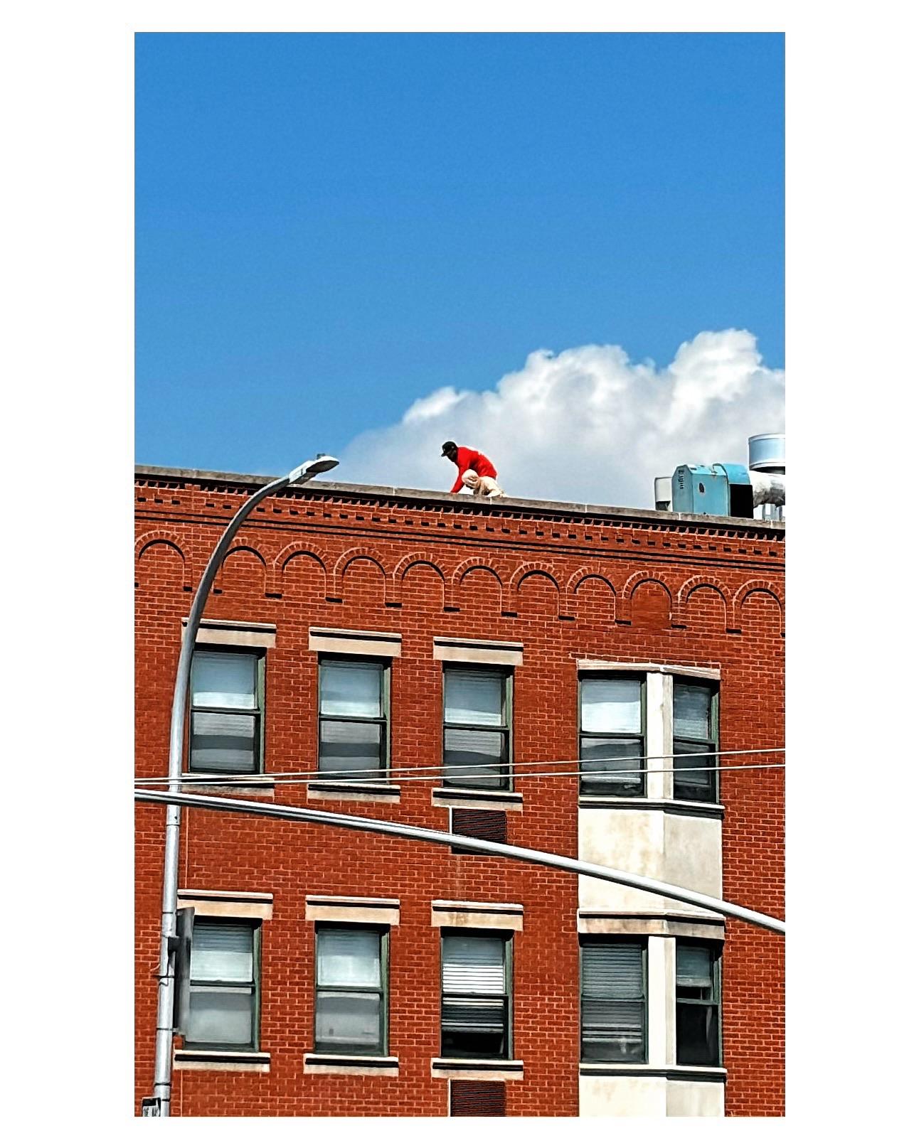 Two workers — one in a pale hoodie, the other in a cap and red sweatshirt — inspect the roofing of a low-rise red brick building. They’re framed by a white cumulus cloud in an otherwise clear blue sky. In front of the building, the horizontal arm of a streetlight crosses the vertical pole of another, creating interesting lines. The image is most striking for its bold colors: the red of the brick and the worker’s shirt, the white cloud, the sky’s vivid blue.