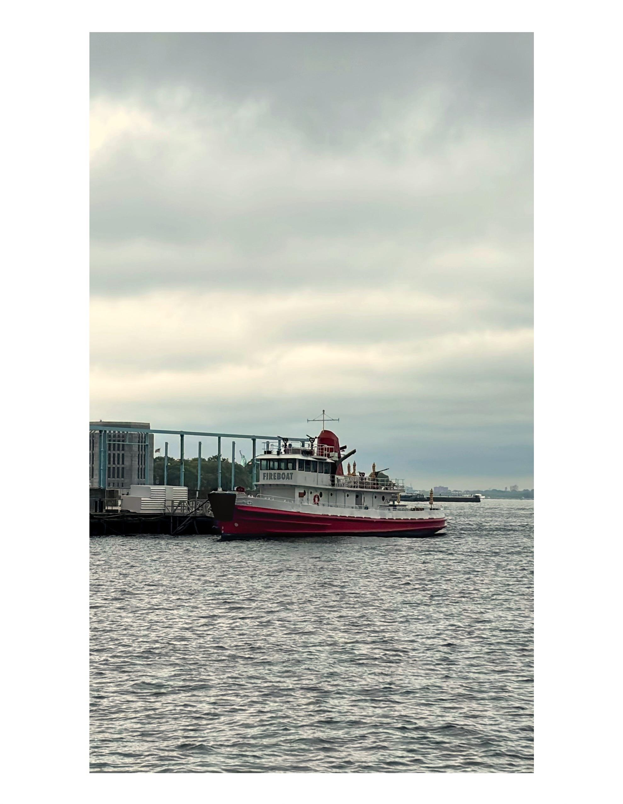 A small ship docked at a pier in New York Harbor, with a red hull and a white deckhouse with “FIREBOAT” painted on the side. In between some vertical metal posts on the pier we can see a building structure and trees on Governors Island, and a tiny Statue of Liberty in the distance. Below is a stretch of calm waves; above is an overcast sky.