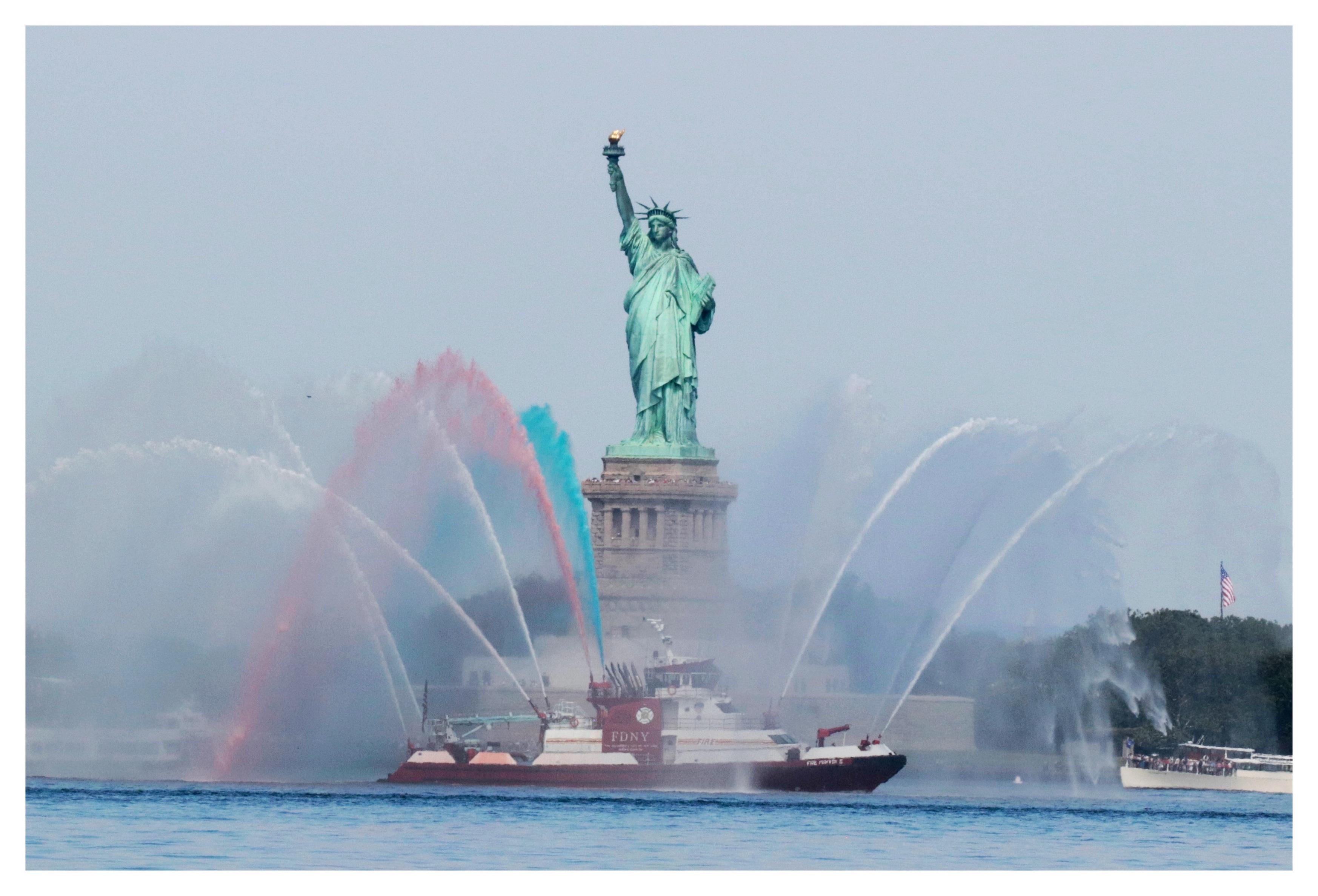 On the waters of New York Harbor, a red-and-white FDNY firefighting ship passes in front of the Statue of Liberty as its hoses spout high arcs of water dyed red, white and blue.