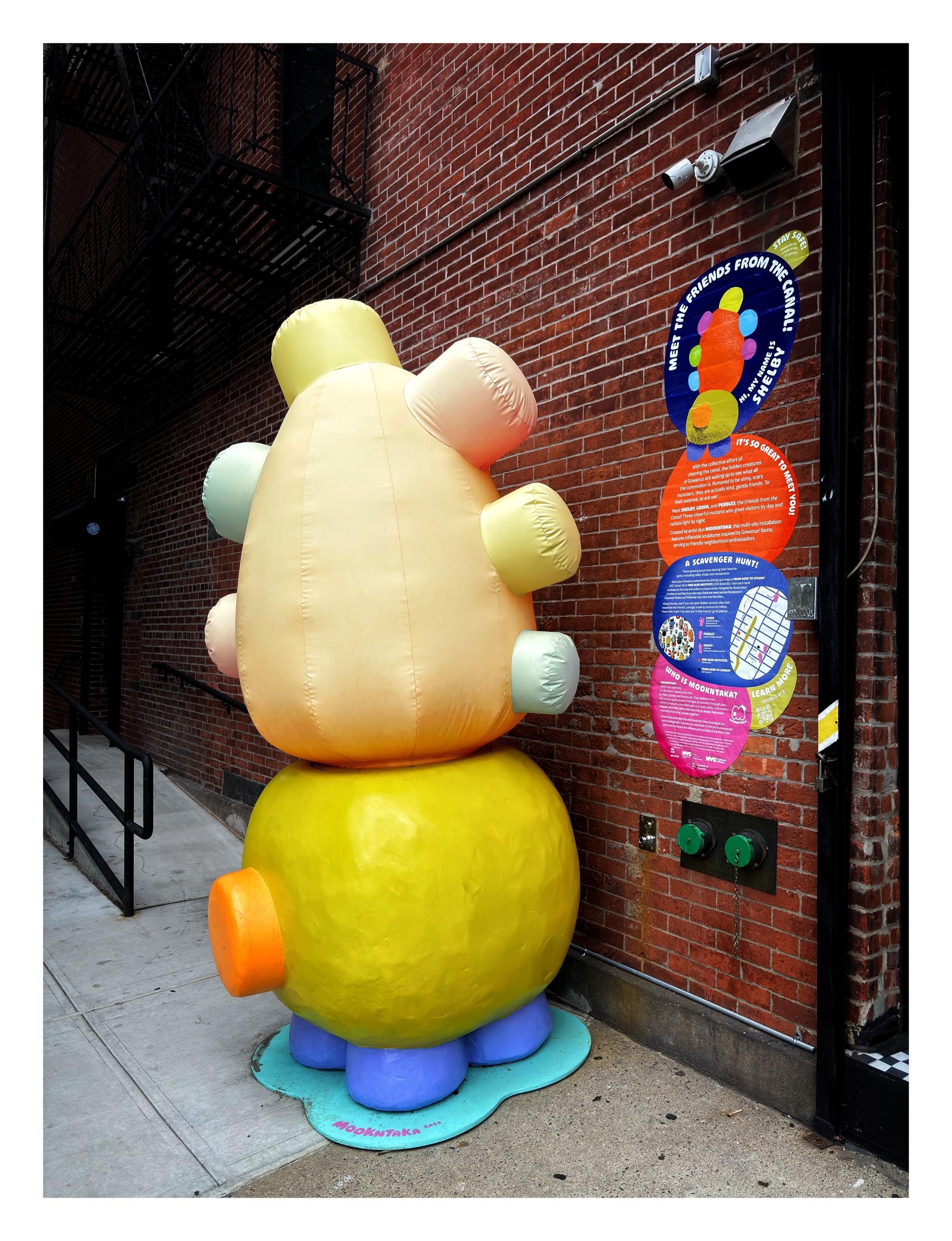 An inflatable public sculpture on a sidewalk outside a brick building. The sculpture is shaped like a faceless creature with “cute” round proportions, with three stumpy legs and a round body and head covered in bumps. The bumps and body parts are in different pastel colors — blue, green, orange, pink — that are supposed to glow at night, though the photo was taken during the day. On the wall beside it is a sticker sign that partially reads:
———
MEET THE FRIENDS OF THE CANAL!

With the collective effort of cleaning the canal, the hidden creatures of Gowanus are waking up to see what all the commotion is. Rumored to be slimy, scary monsters, they are actually kind, gentle friends. To their surprise, so are we!

Meet SHELBY, LOOFA, and PEBBLES, the Friends from the Canal! Three cheerful mutants who greet visitors by day and radiate light by night.

Created by artist duo MOOKNTAKA, this multi-site installation features inflatable sculptures inspired by Gowanus’ fauna, serving as friendy ne