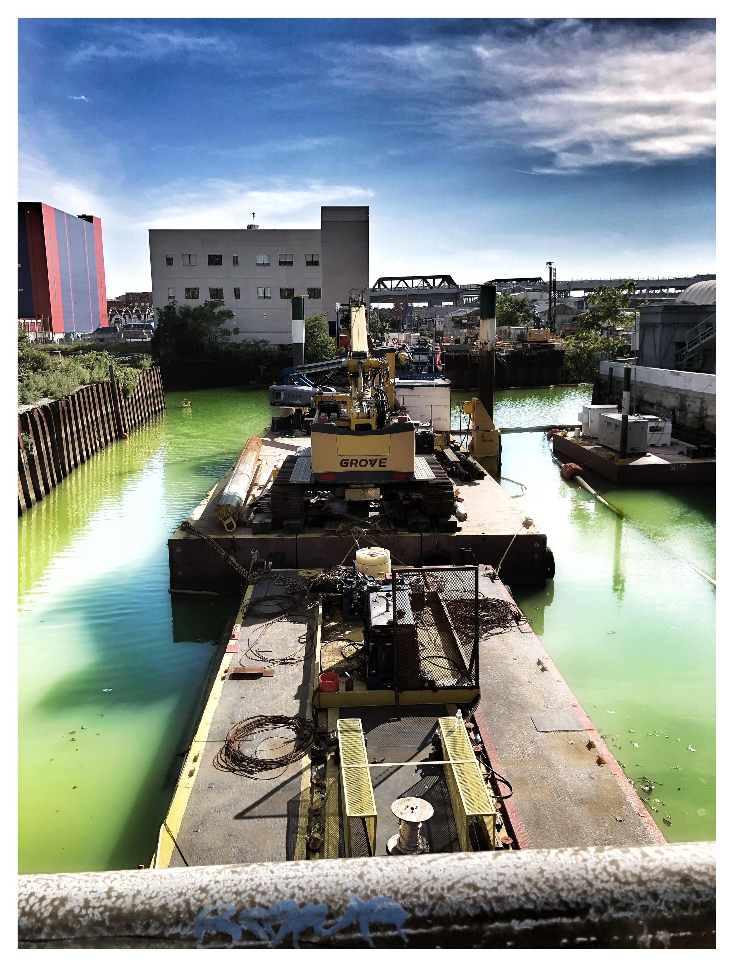 A view of Brooklyn’s Gowanus Canal as seen from behind the rusted railing of a pedestrian overpass. The canal recedes from camera and curves to the right, surrounded by an industrial landscape of buildings and bridges under a blue sky with clouds. Its famously toxic water is a sickly green, divided into two channels by floating barges of dredging equipment — part of the effort to clean up the canal by scooping out the “black mayonnaise” at its bottom (a sludge of “oil, coal, pesticides, rotting debris, raw sewage, chemicals, and heavy metals, which includes arsenic, benzene, chromium, mercury, and lead,” according to Gothamist).

More lovely details from Gothamist: “The canal’s waters are tainted by millions of gallons of sewage every year, which, in the 1970s, led to it being contaminated with typhoid, cholera and tuberculosis. In more recent years, it has been diagnosed with gonorrhea. The canal’s coastline has been poisoned by a century of industrial use, including chemical factorie